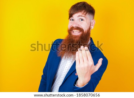 Young handsome red haired businessman standing against yellow background, inviting to come with hand. Happy that you came