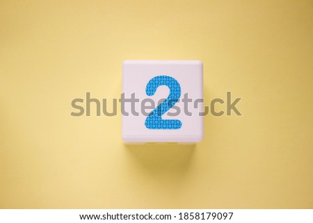 Close-up photo of a white plastic cube with a blue number 2 on a yellow background. Object in the center of the photo