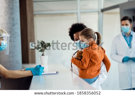 African American female doctor and small girl talking to a nurse at reception desk at medical clinic while wearing protective face masks. 