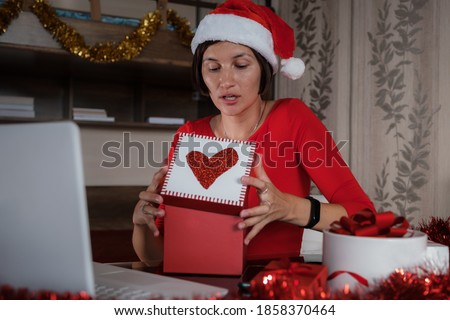 Remote Christmas video party. Family quality time during pandemic. Online video call conferencing. Young woman wearing red Santa hat and holding gift box. Virtual meeting conference calling from home