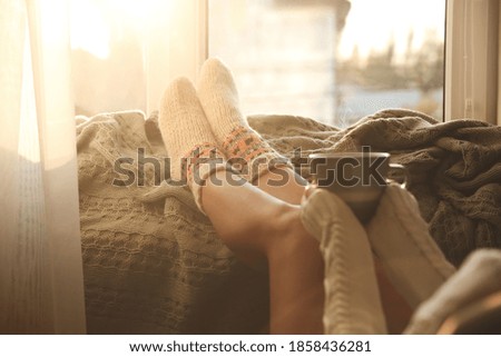 Woman in knitted socks holding cup of hot drink on plaid near window at home, closeup