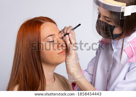 A makeup artist in a robe, mask and gloves makes makeup for a red-haired girl.