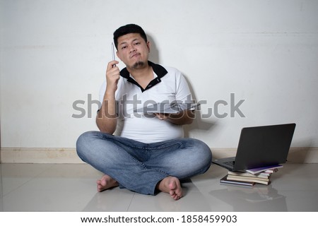 Portrait of a happy young man in white Polo-shirt holding laptop computer while sitting on a floor isolated over white background