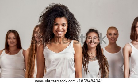 Portrait of young african american woman in white shirt smiling at camera. Group of diverse women standing isolated over grey background. Selective focus. Front view. Web Banner Royalty-Free Stock Photo #1858484941