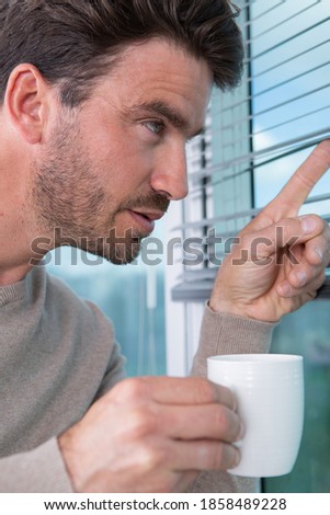 man holding a coffee and looking through window blind