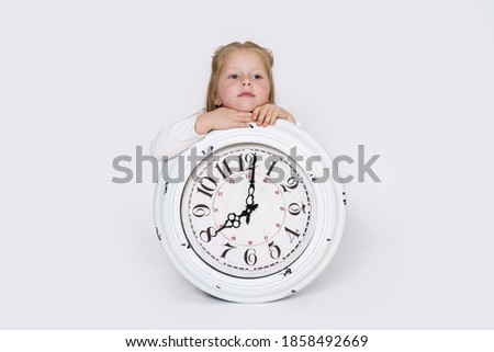 Children concept. The girl is sitting on the floor in front of a large white clock. Isolated over white background.