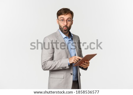 Image of surprised businessman with beard, wearing grey suit and glasses, saying wow and looking impressed after reading something on digital tablet, standing over white background