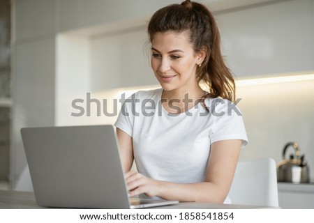 a woman is typing sitting at home in the kitchen at a laptop computer doing tasks to work online in quarantine. portrait of brunette hair in a ponytail beautiful in a comfortable white t-shirt.