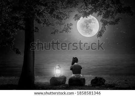 A black and white picture of a young woman sitting on a log with a lamp by the sea, looking at the full moon.