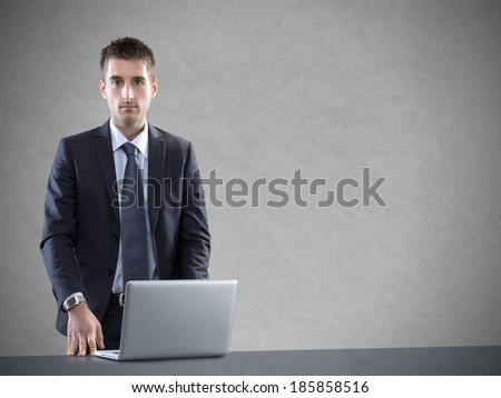 Young businessman with laptop leaning on desk against gray background.