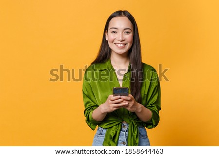 Smiling cheerful young brunette asian woman wearing basic green shirt standing using mobile cell phone typing sms message looking camera isolated on bright yellow colour background, studio portrait