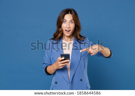 Shocked young brunette woman 20s wearing basic jacket standing pointing index finger on mobile cell phone typing sms message looking camera isolated on bright blue colour background, studio portrait