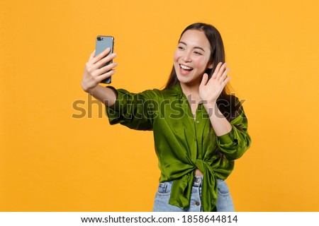 Excited joyful young brunette asian woman wearing basic green shirt standing doing selfie shot on mobile phone waving greeting with hand isolated on bright yellow colour background, studio portrait