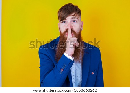 young red haired bearded businessman standing against yellow background  makes silence gesture, keeps finger over lips. Silence and secret concept.