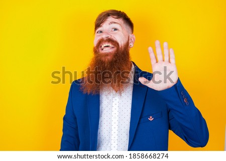 young red haired bearded businessman standing against yellow background Waiving saying hello happy and smiling, friendly welcome gesture.