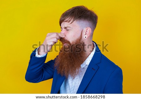 young red haired bearded businessman standing against yellow background smelling something stinky and disgusting, intolerable smell, holding breath with fingers on nose. Bad smell