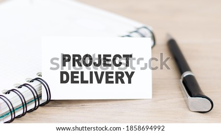 text PROJECT DELIVERY on a white card, which is next to the notebook and pen on the table.