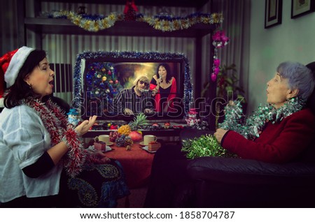 A professional caregiver and a senior adult woman sitting in front of a TV on Christmas Eve. They sing carols along with singers on the holiday channel. The room is decorated with tinsels.