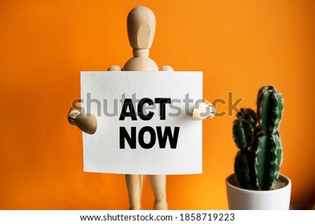 The wooden man holds a white sign with the text ACT NOW in his hands. The content of the lettering has implications for business concept and marketing.