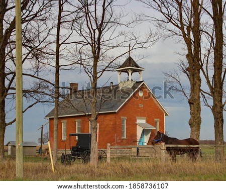 Bare winter trees with horse and buggy in the foreground and old red brick school in the background of this rural scene.  Royalty-Free Stock Photo #1858736107