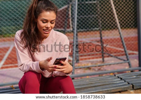 Close up photo of a sportswoman sitting and resting after workout, using mobile phone, smiling and contemplating view