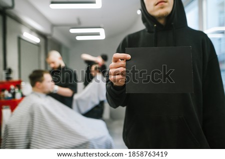 A man in a black hoodie holds a black blank card in his hand on a background of barbers cutting clients in a bright men's hairdresser. Barber holds a place for copy space, standing in a barbershop.