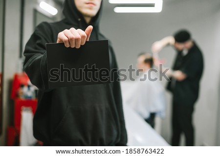 Barber in dark hoodie holds a card for copyspace on the background of a male hairdresser and barber clipping clients. Man holding blank black card, focus on blank space on barbershop background