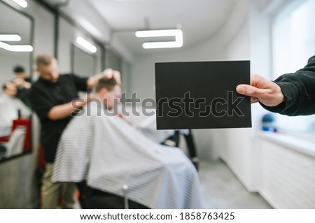 Male hand holding blank black card for copyspace on background of male hairdresser and barber clipping clients. Barber holds a dark blank card in his hand on the background of a male hairdresser