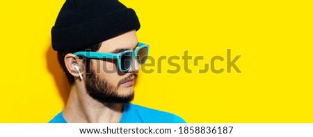 Close-up studio portrait of young man using wireless earbuds or earphones. Yellow background with copy space. Wearing blue sunglasses and black hat.