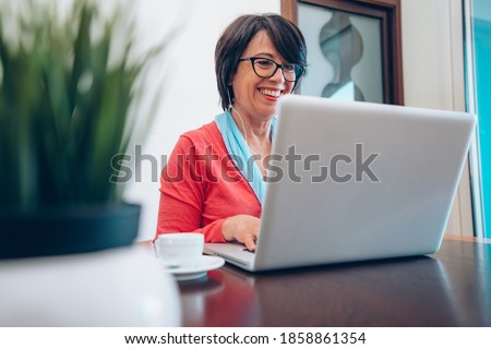 Mature woman using pc laptop indoor - Senior female smart working with computer at home - She is sitting on a wooden modern desk at home. Smiling and enjoying communication