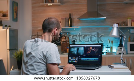 Freelancer editing a video using post production software working from home overtime sitting in the kitchen. Videographer processing audio film montage on professional laptop late at night