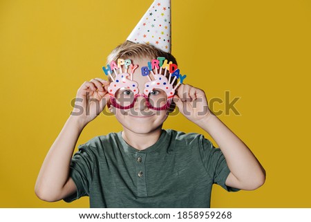 Portrait of a cute smiling ten year old birthday boy standing in front of a yellow wall. He is wearing birthday glasses and party cone. Posing for a photo.