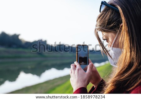 young woman in a red coat wearing a medical mask while photographing a river bank with her phone