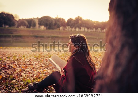 young woman with red coat reading a book sitting on the ground in an autumn park surrounded by yellow leaves