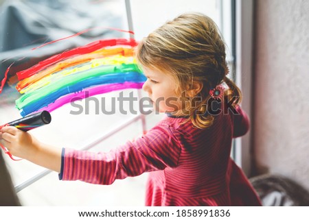 Adorable little toddler girl with rainbow and heart painted with colorful window color during pandemic coronavirus quarantine. Child painting rainbows around the world with the words Let's all be well