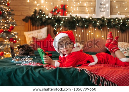 Cute boy in Santa Claus costume watching Christmas cartoons on tablet, preparing for celebration, wonderful atmosphere and decorations, miracle