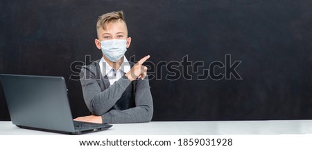 Teen boy wearing protective face mask sits with laptop at school and points away on empty blackboard during corona virus and flu outbreak. Empty space for text