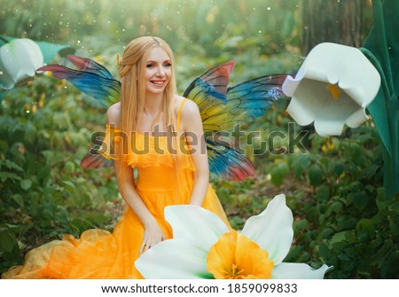 Beautiful happy woman fairy nymph sitting on forest. Magical fantasy wings costume pixie butterflies. Elf girl princess long yellow dress. blond hair smiling face. Summer nature tree, green grass.
