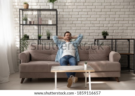 Peaceful young man relaxing meditating on comfortable sofa with folded hands behind hand, breathing fresh air, daydreaming napping enjoying calm mindful lazy weekend moment alone in modern living room Royalty-Free Stock Photo #1859116657