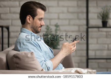 Addicted to technology young relaxed man in eyeglasses sitting on comfortable couch, spending time online communicating with friends in social network, playing mobile games or using applications.