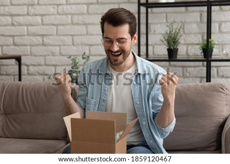 Emotional happy young man in eyeglasses opening small cardboard parcel, celebrating getting wished item, satisfied with shopping experience in online store or fast delivery service, getting gift. Royalty-Free Stock Photo #1859120467
