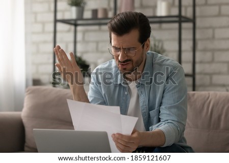 Frustrated young man in glasses looking at paper documents stressed by mistake, bank debt notification or eviction notice, having financial problems or unpaid bills, shocked by unexpected news. Royalty-Free Stock Photo #1859121607