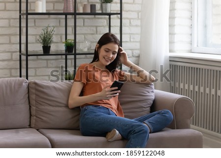 Carefree young attractive woman resting on comfortable couch, web surfing internet information, using smartphone dating application, reading social media, spending weekend time online at home.