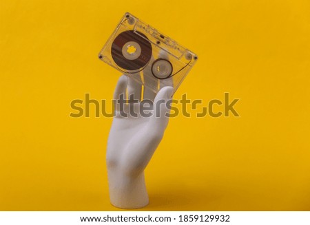 White mannequin hand holding retro audio cassette on yellow background.