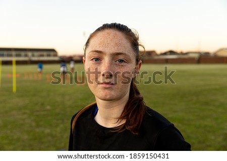 Portrait close up of a young adult Caucasian female rugby player standing on a rugby pitch looking to camera, with her teammates training in the background Royalty-Free Stock Photo #1859150431