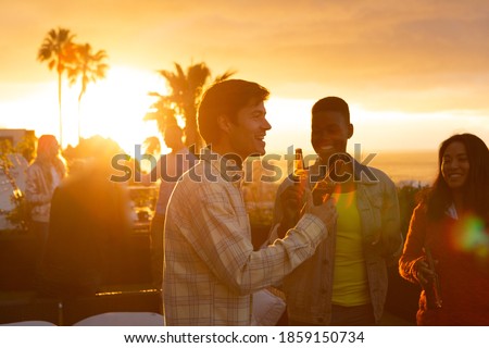 Front view of a multi-ethnic group of friends hanging out on a roof terrace with a sunset sky, holding bottles of beer and smiling Royalty-Free Stock Photo #1859150734
