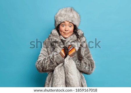 Winter and Christmas concept. Surprised Asian woman with two pigtails dressed in winter fur hat coat shocked to see heavy snowfall outside poses against blue background. Arctic female in warm clothing