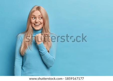 Positive blonde smiling young European woman points thumb away at blank copy space demonstrates advert dressed in casual turtleneck isolated over blue background. Look here and follow this way