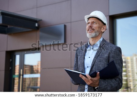 Monitoring the process. Confident middle aged architect, engineer manager wearing helmet looking away while standing outdoors and holding a blueprint