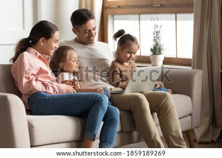 Beautiful full family with two cute daughters resting with laptop seated on sofa. Couple develop offspring watching educational online tv, buy goods on internet, have fun use modern technology concept